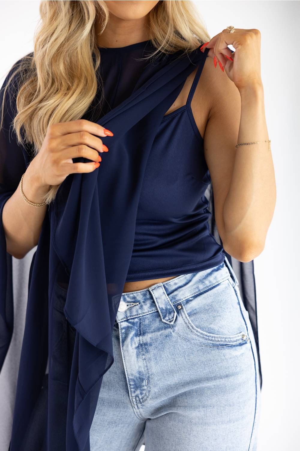 A woman wearing a navy blue sleeveless top with cutout details and a matching sheer draped cardigan, paired with light-wash blue jeans.