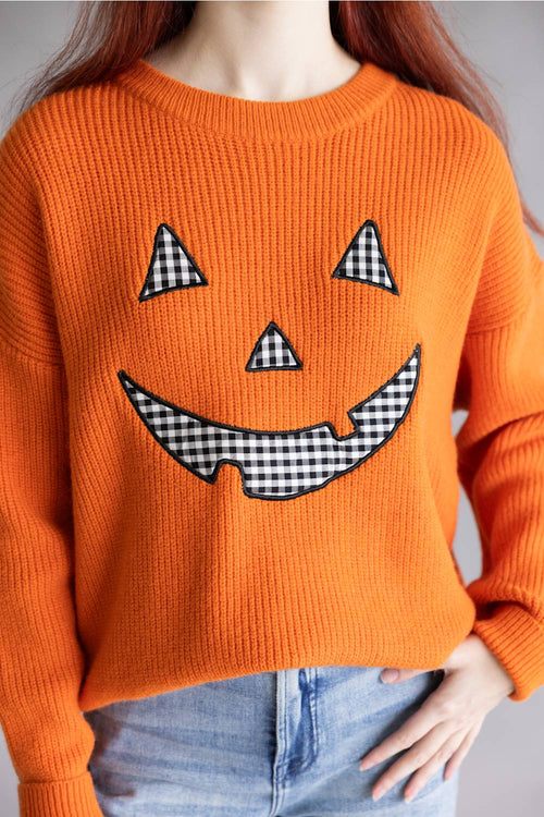 An orange crewneck knit sweater featuring a large jack-o'-lantern face design with black and white gingham patterned details, worn by a model with red hair and blue jeans.