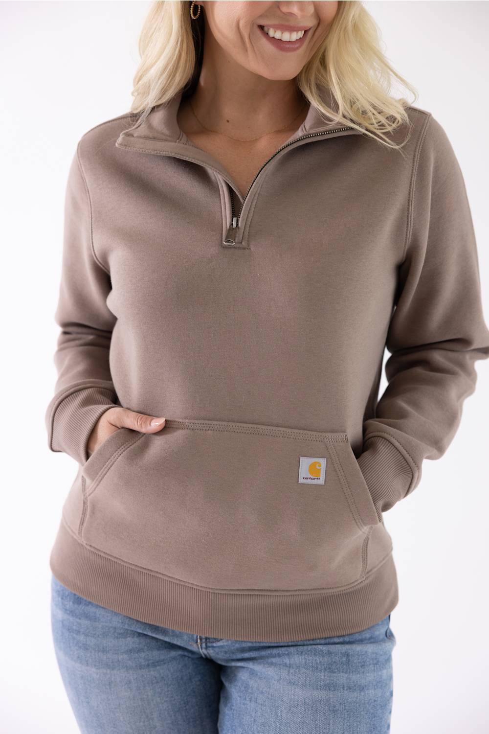 Front view of a stone brown Carhartt quarter-zip sweatshirt featuring a mock-neck collar and a front kangaroo pocket with a small logo patch, worn with blue jeans.