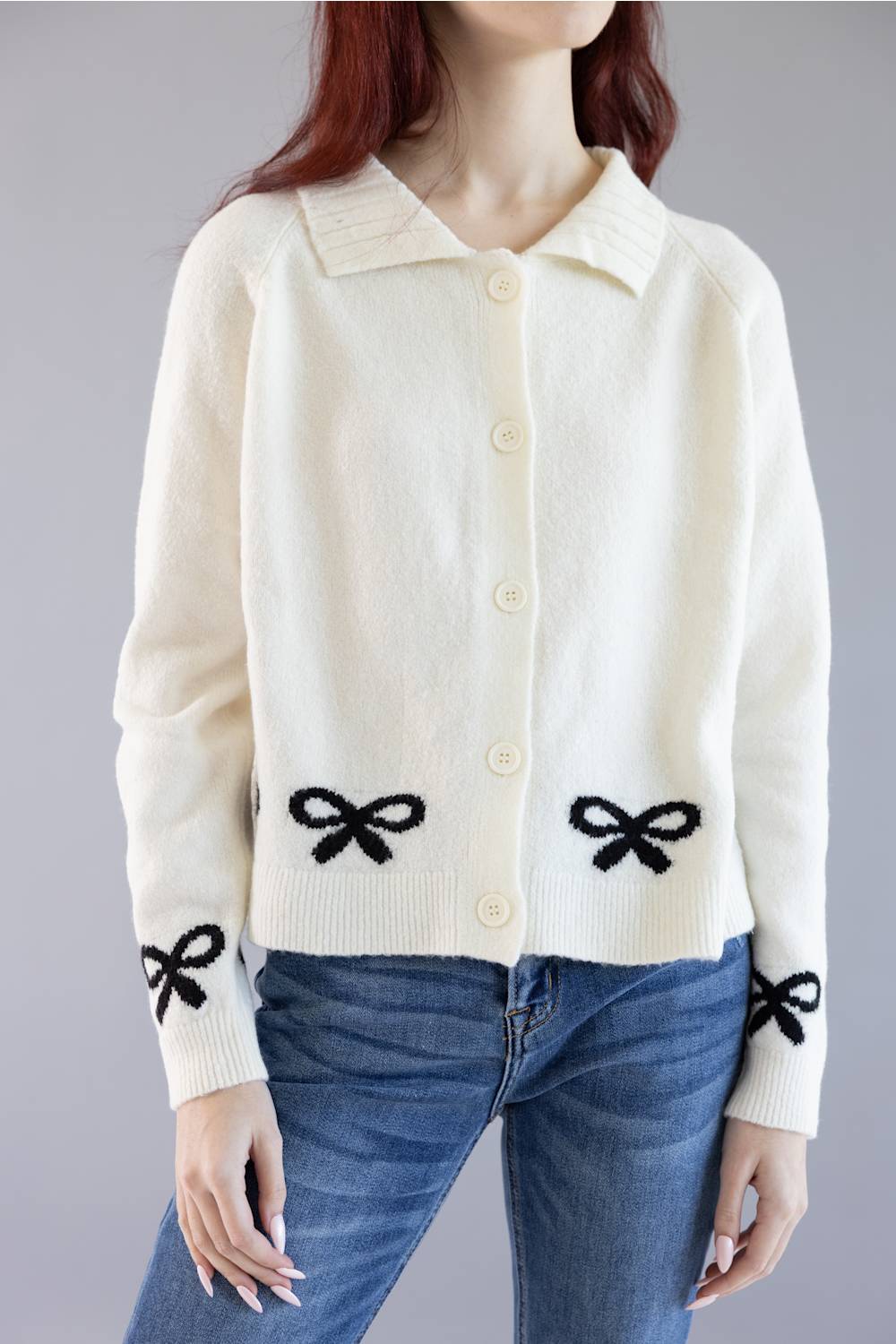 An off-white knit cardigan sweater with black knitted bow details on the front and cuffs, worn by a model with blue jeans against a neutral gray background.