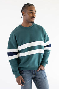 A man viewed from behind wearing a teal colorblock hoodie with white and navy sleeve stripes, paired with light-wash blue jeans, against a plain white background.