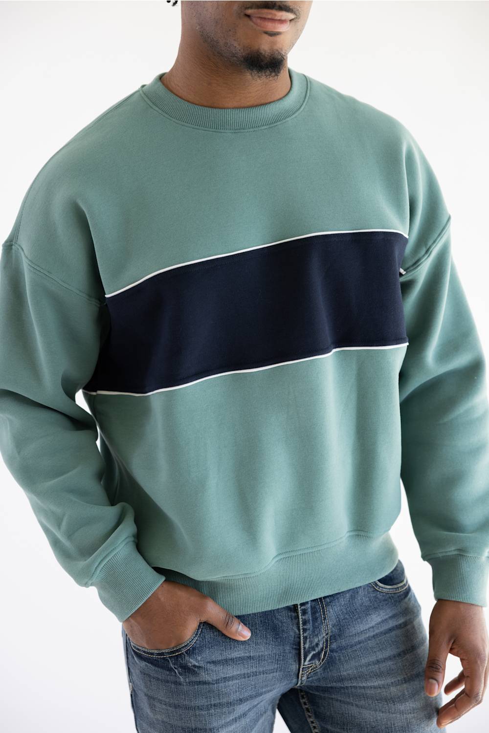 A man wearing a sage green crewneck sweatshirt with a wide navy blue horizontal stripe across the chest, paired with blue jeans, against a plain white background.