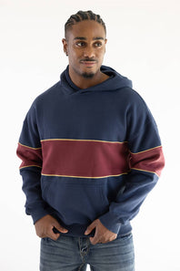 A man wearing a navy blue hoodie with a wide burgundy horizontal stripe across the chest and thin gold trim, paired with blue jeans, against a plain white background.