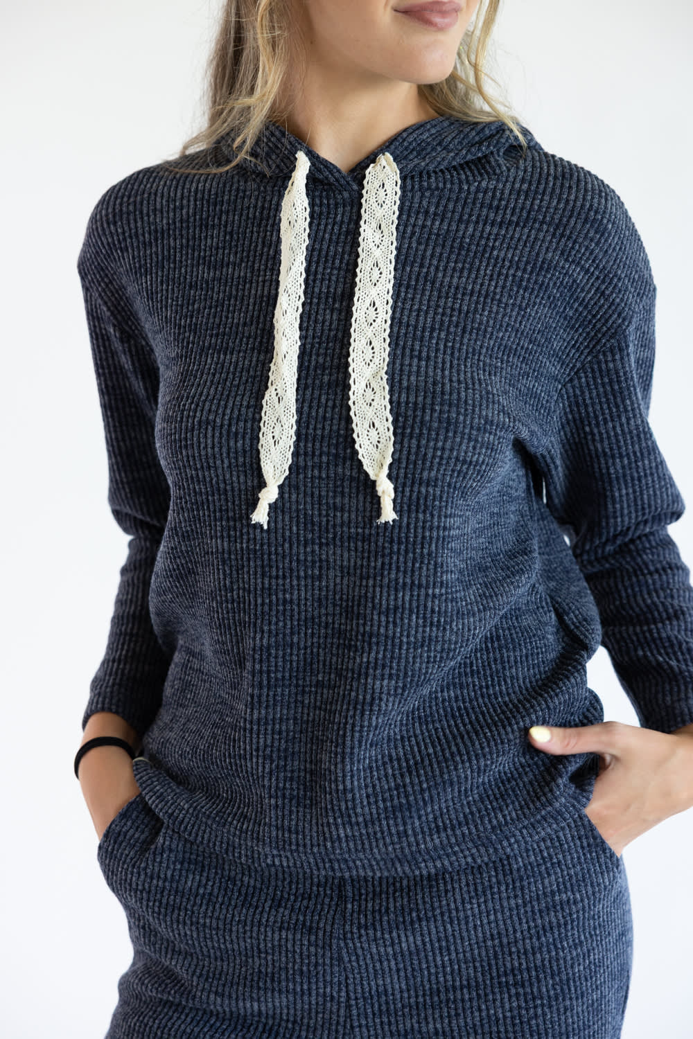 A woman wearing a navy blue corduroy hoodie with cream-colored lace drawstrings, shown from the chest down against a plain white background.