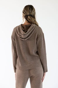 A woman viewed from the back wearing a ribbed knit hoodie and matching pants in a sand-brown color, set against a plain white studio background.