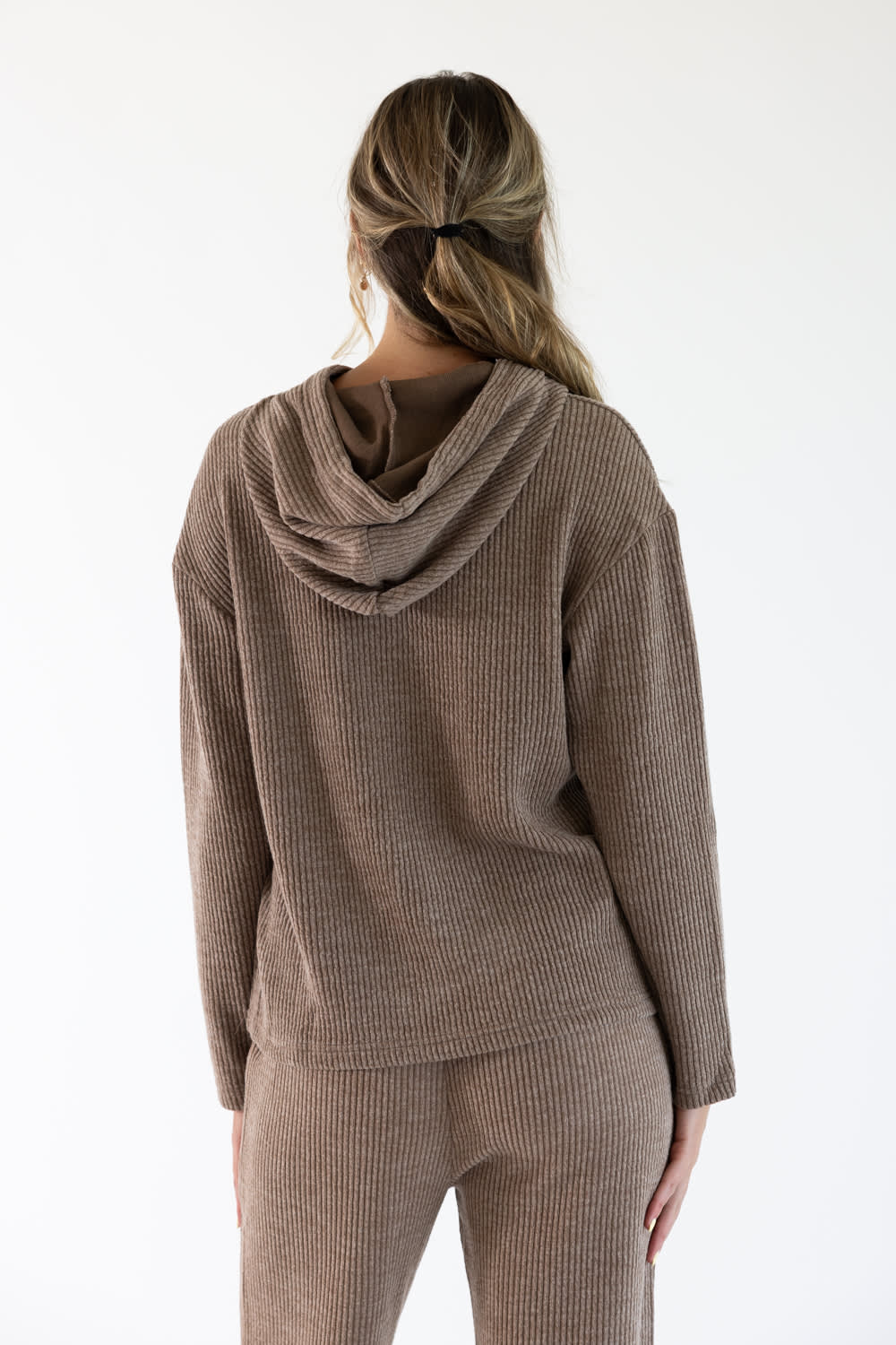 A woman viewed from the back wearing a ribbed knit hoodie and matching pants in a sand-brown color, set against a plain white studio background.