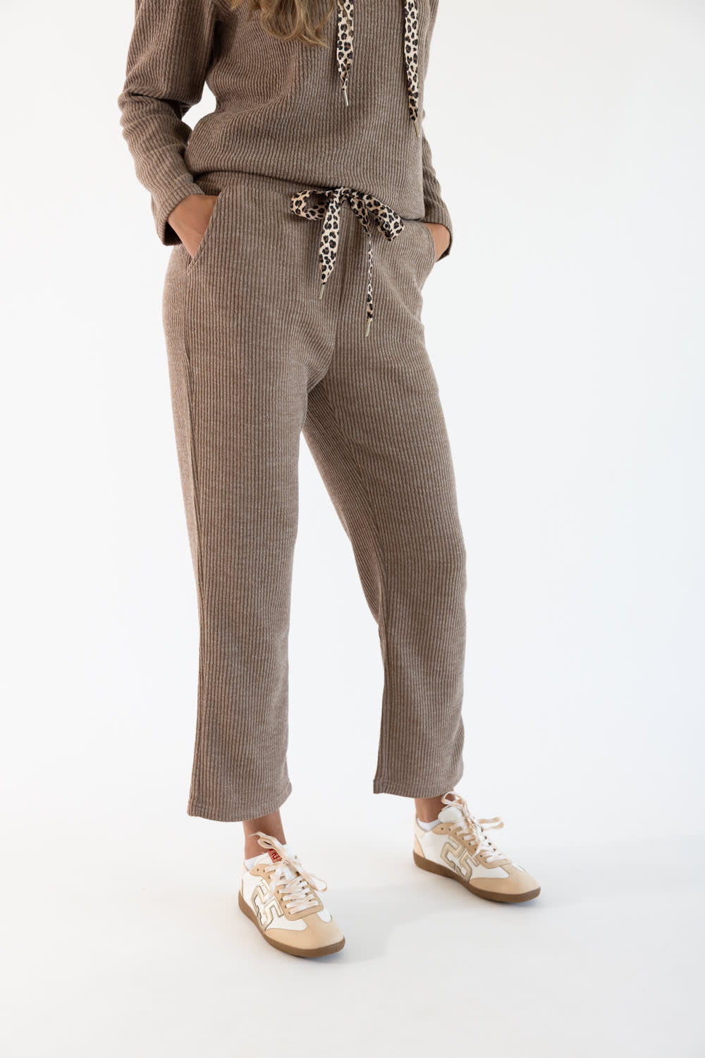 A model wearing a matching corduroy lounge set consisting of a long-sleeve top and lounge pants in sand brown, featuring a leopard-print drawstring and matching leopard-print hood, paired with beige sneakers.