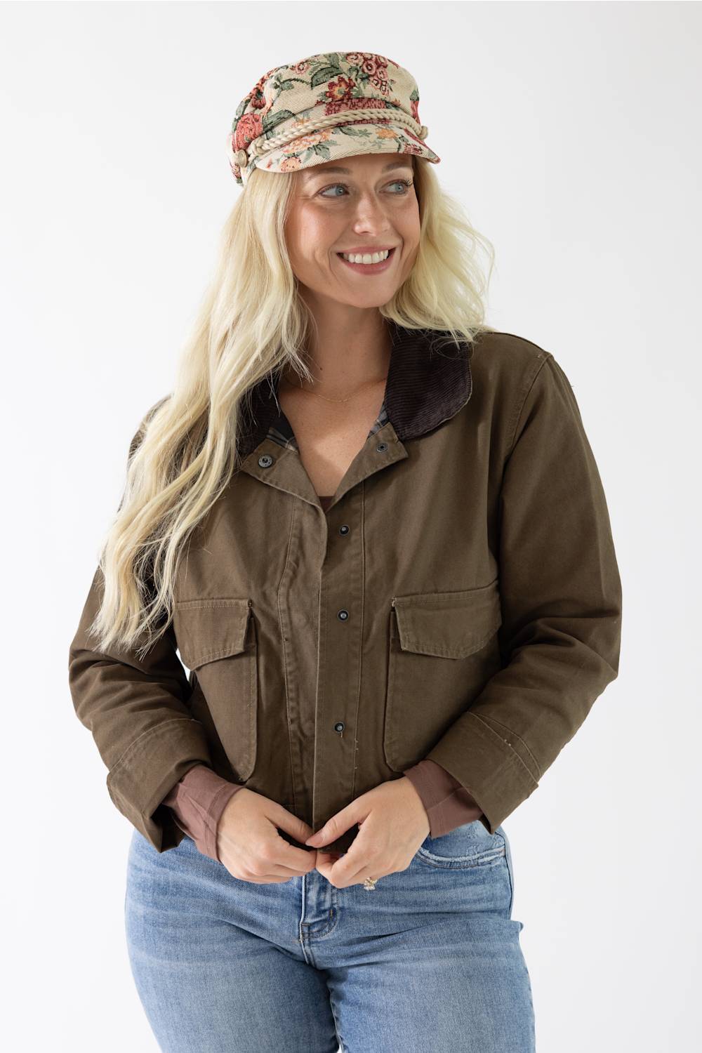 Woman wearing an olive green canvas jacket with a dark brown corduroy collar, styled with a floral cap and blue jeans.