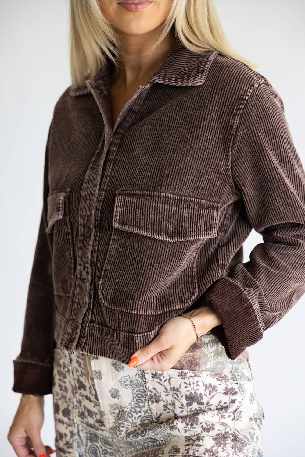 A woman wearing a dark brown corduroy jacket with two front flap pockets, paired with a beige and brown patterned skirt, against a plain light gray background.