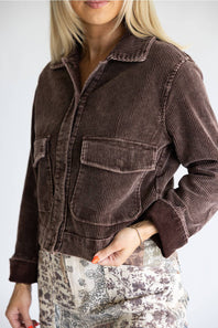 A woman wearing a dark brown corduroy jacket with two front flap pockets, paired with a beige and brown patterned skirt, against a plain light gray background.