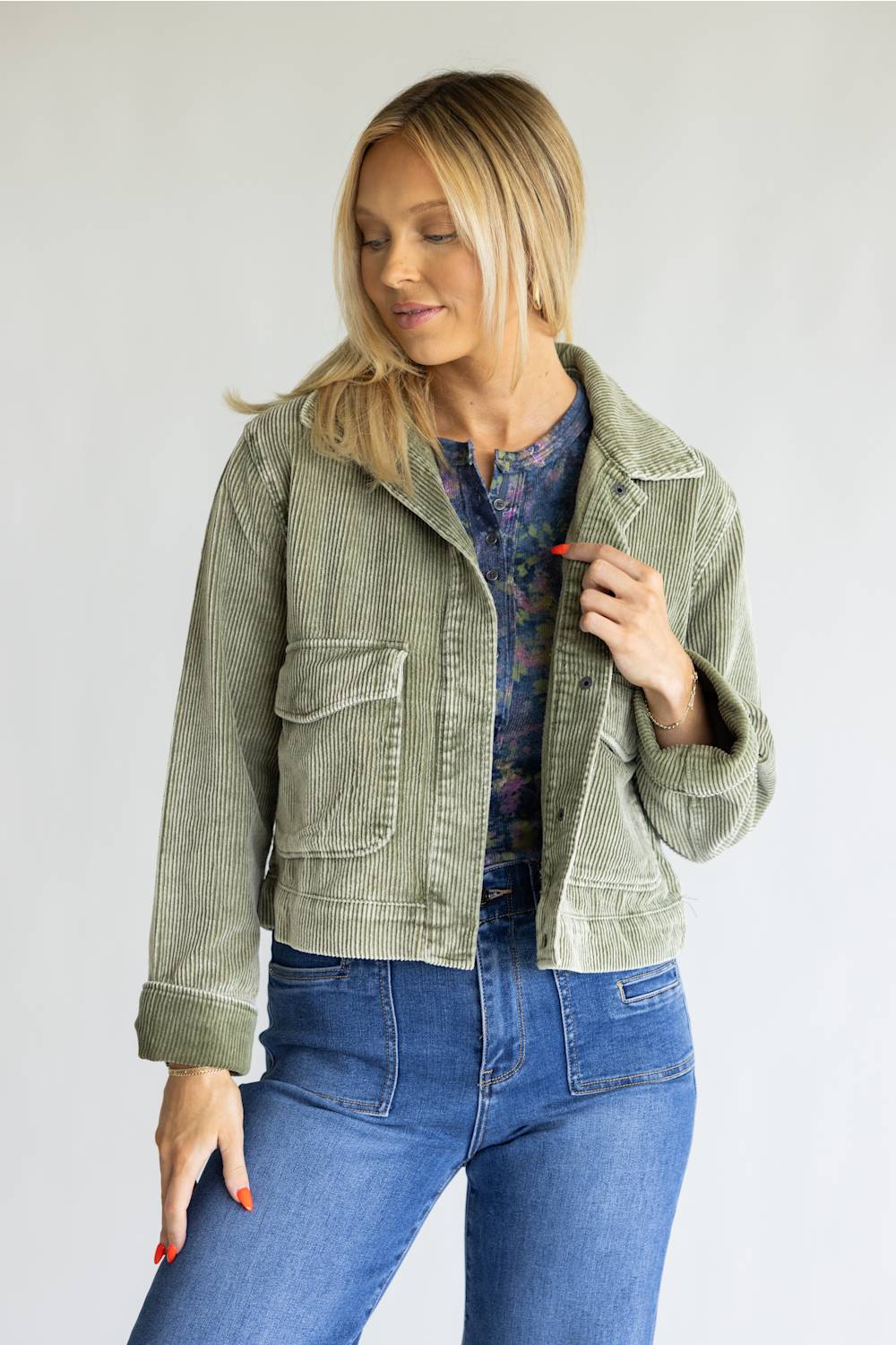 A woman wearing a light sage green corduroy jacket over a blue floral top and blue jeans, standing against a plain light gray background.