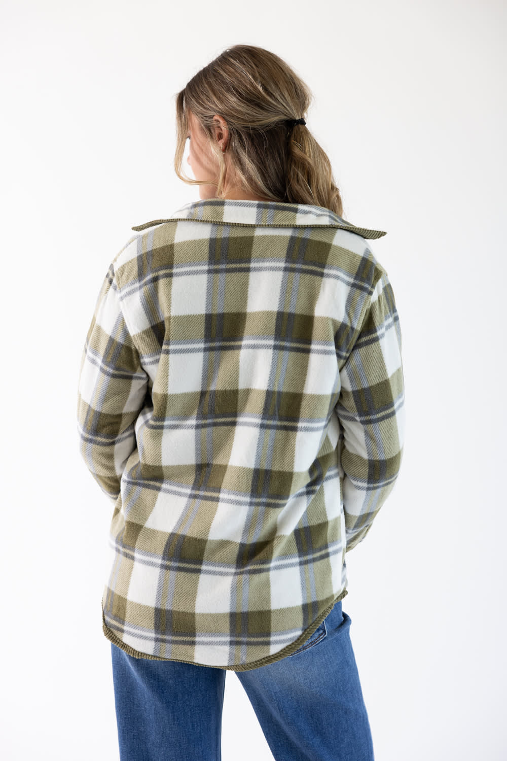 A woman viewed from behind wearing a long-sleeve plaid flannel shirt in olive green, white, and blue, paired with blue jeans, against a plain white background.