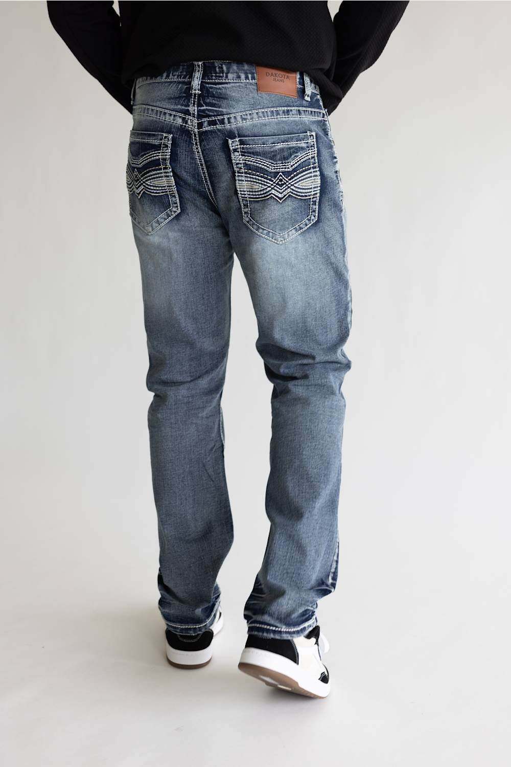 Rear view of a person wearing light blue denim jeans with decorative stitching on the back pockets, a black top, and black-and-white sneakers against a plain light gray studio background.