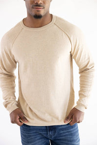 A man wearing a light beige knit crewneck sweater with raglan sleeves, paired with blue jeans, against a plain white background.