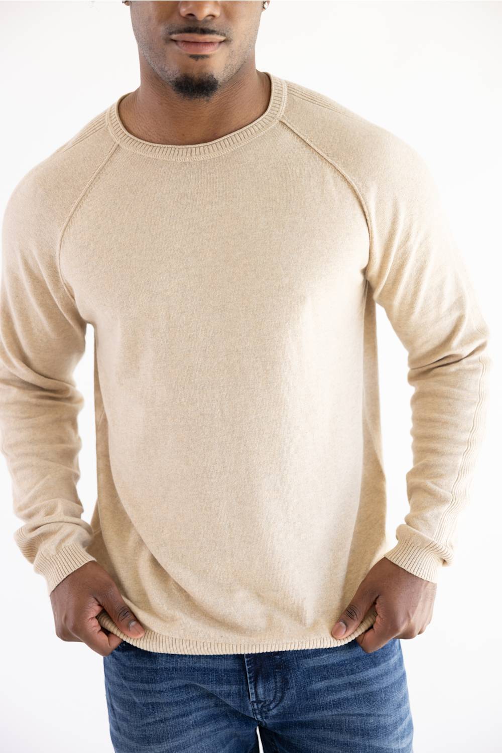 A man wearing a light beige knit crewneck sweater with raglan sleeves, paired with blue jeans, against a plain white background.