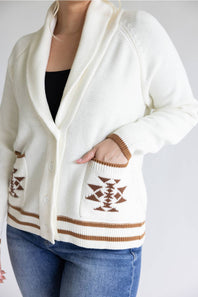 A close-up of a person wearing a cream knit cardigan with brown geometric patterns on the pockets and striped trim, paired with a black top and blue jeans.