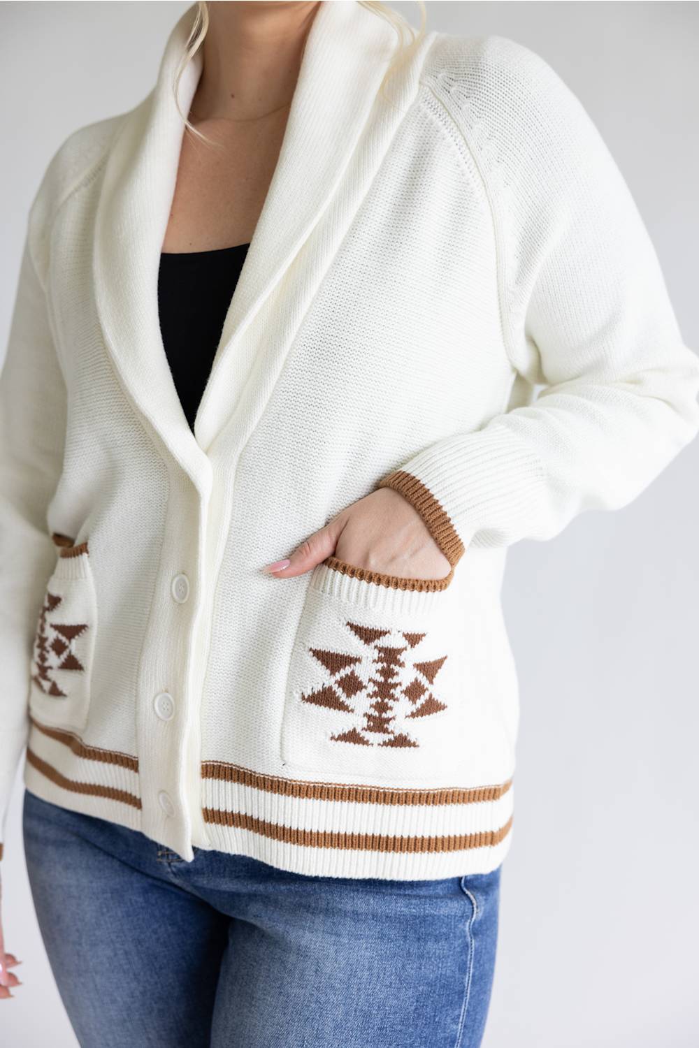 A close-up of a person wearing a cream knit cardigan with brown geometric patterns on the pockets and striped trim, paired with a black top and blue jeans.