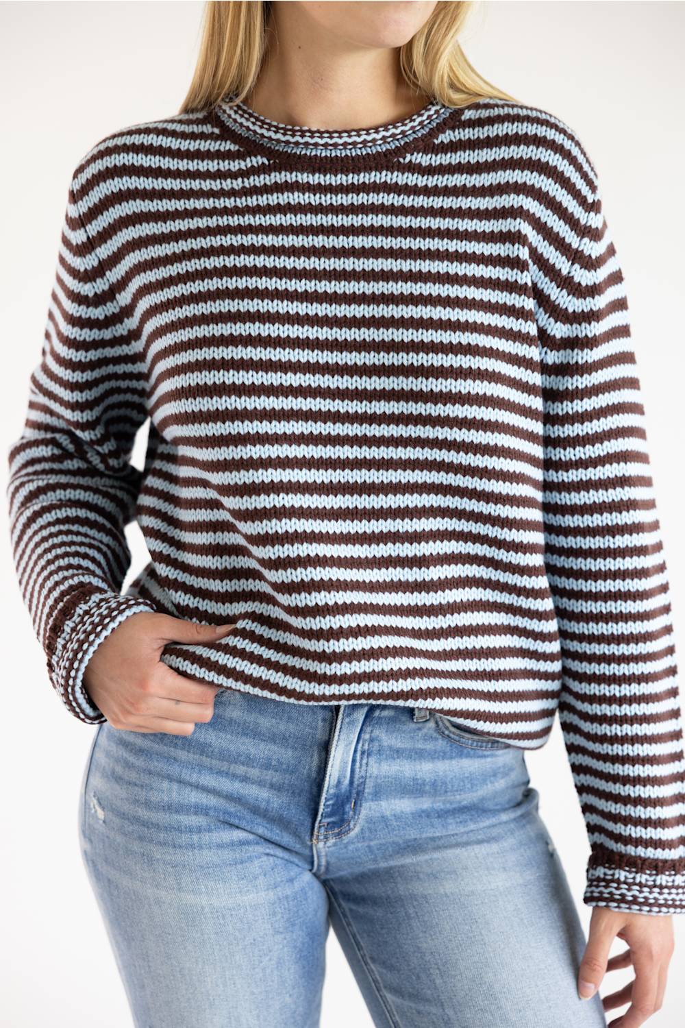 A woman wearing a long-sleeve knit sweater with horizontal brown, white, and light blue stripes, paired with blue jeans against a plain white background.