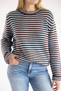 A woman wearing a long-sleeve knit sweater with horizontal brown, white, and light blue stripes, paired with blue jeans against a plain white background.