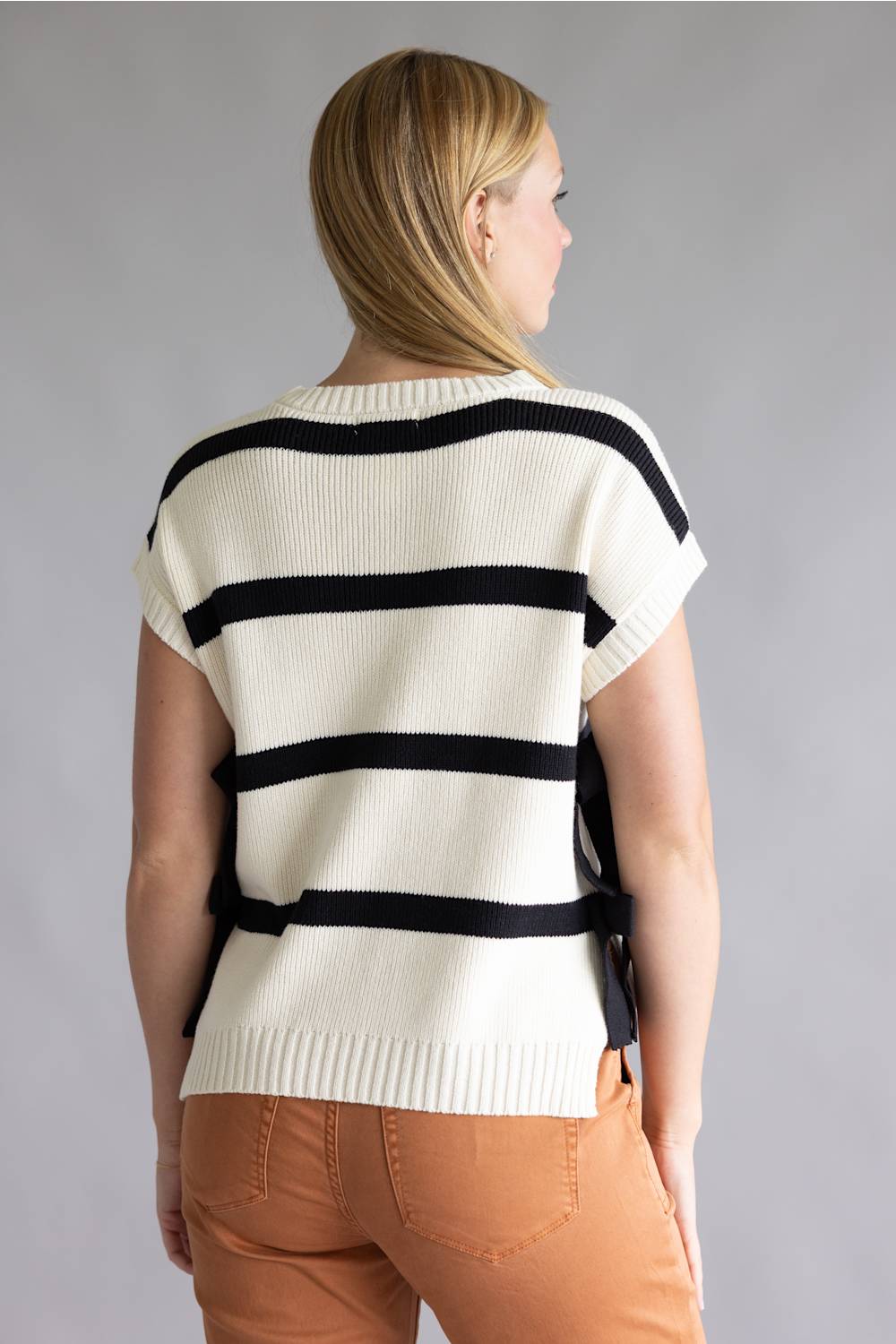A woman shown from the back wearing a cream knit sweater with bold black horizontal stripes and peach-colored pants, set against a plain gray studio background.