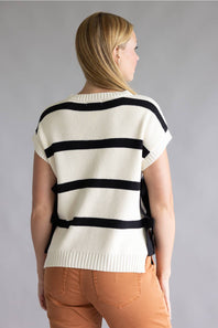 A woman shown from the back wearing a cream knit sweater with bold black horizontal stripes and peach-colored pants, set against a plain gray studio background.