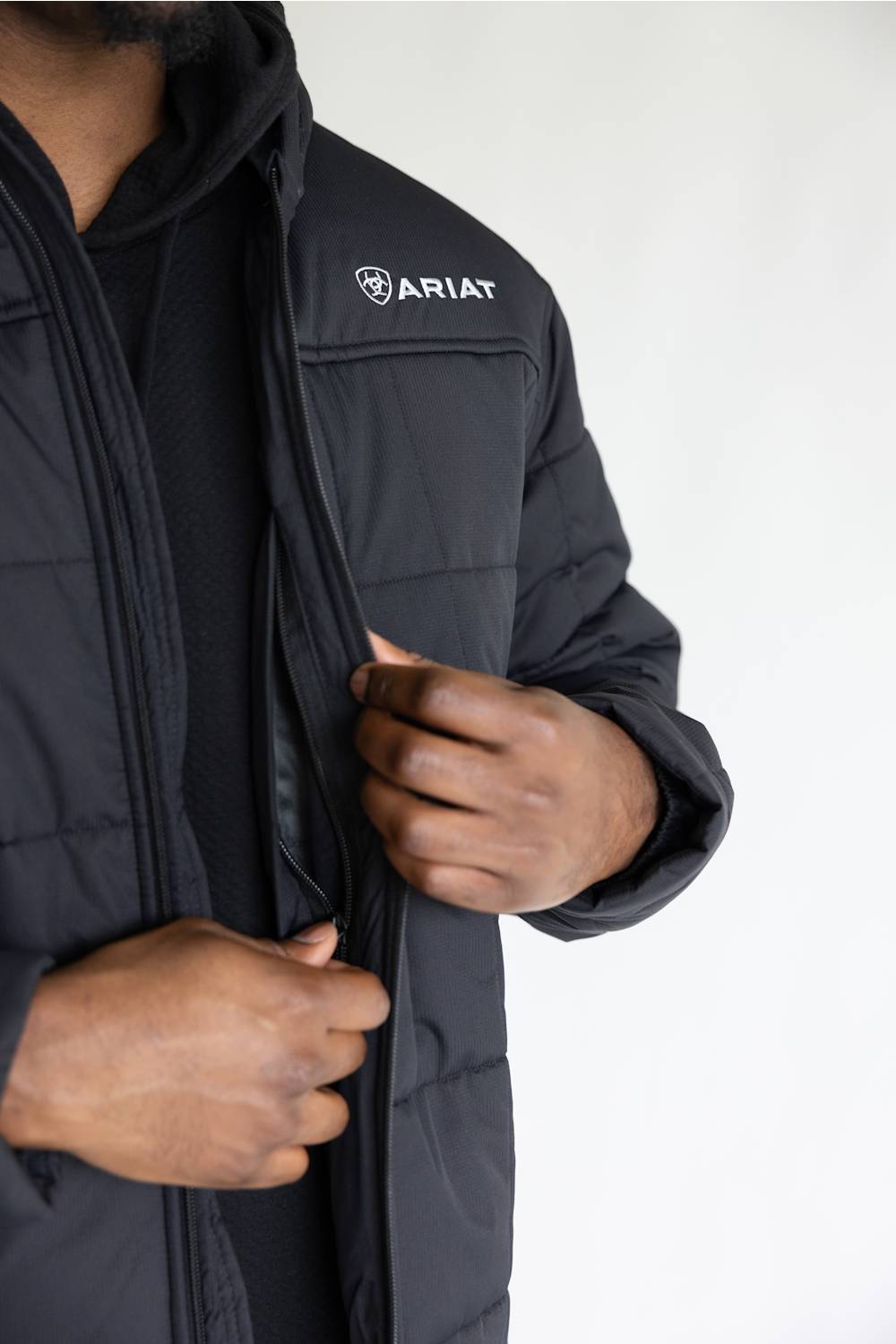 A close-up of a person wearing a black Ariat puffer jacket over a black hoodie, with the person's hands opening the front zipper of the jacket against a plain white background.