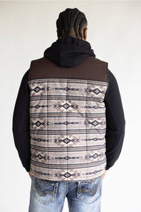 A man viewed from behind wearing a brown hooded pullover vest with a beige, black, and brown Southwestern geometric patterned back panel, paired with a black hoodie and blue jeans.