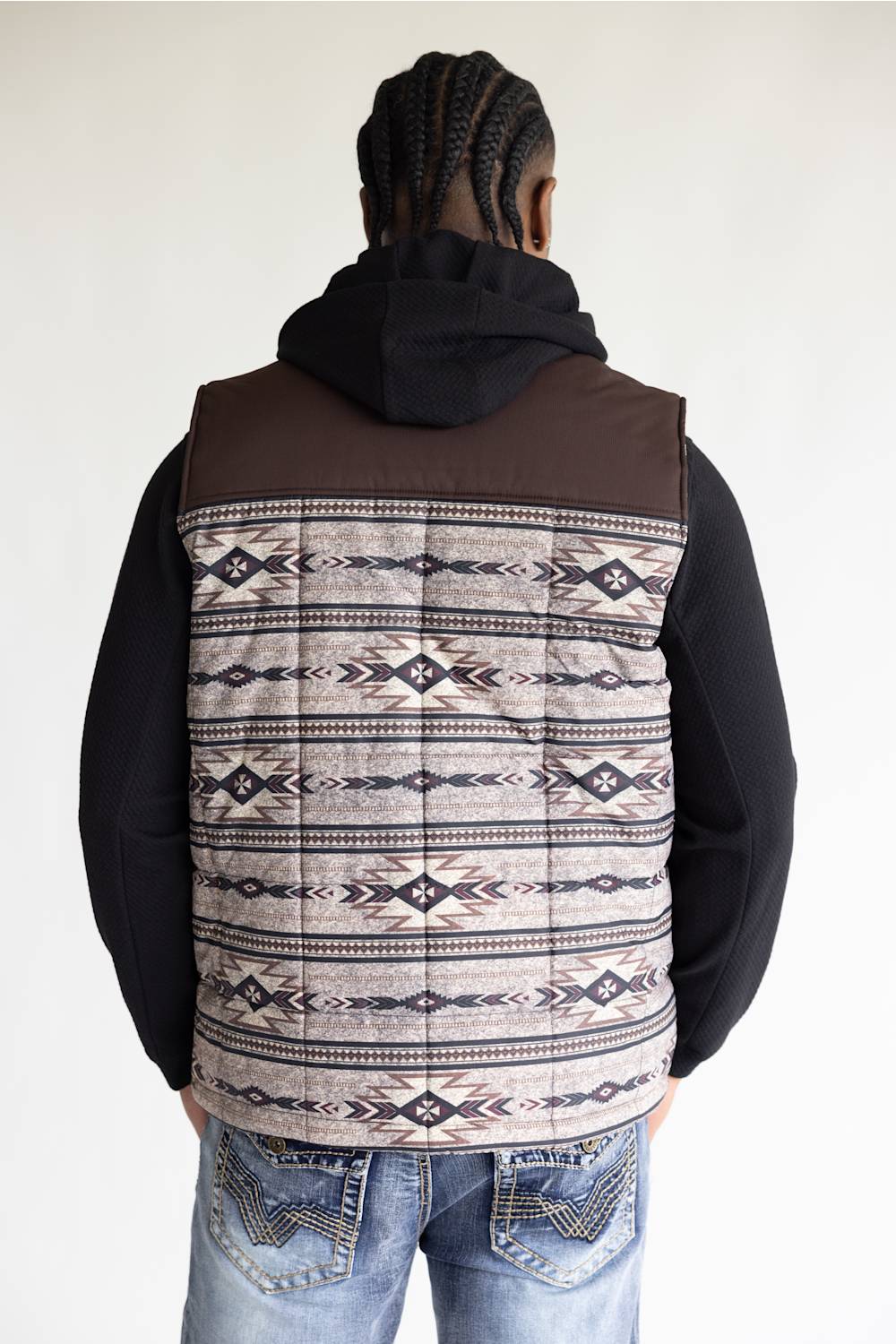 A man viewed from behind wearing a brown hooded pullover vest with a beige, black, and brown Southwestern geometric patterned back panel, paired with a black hoodie and blue jeans.
