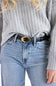 A close-up of a person wearing a black -leather belt with a large oval gold-tone buckle, styled with a gray ribbed knit sweater and light-wash blue jeans.
