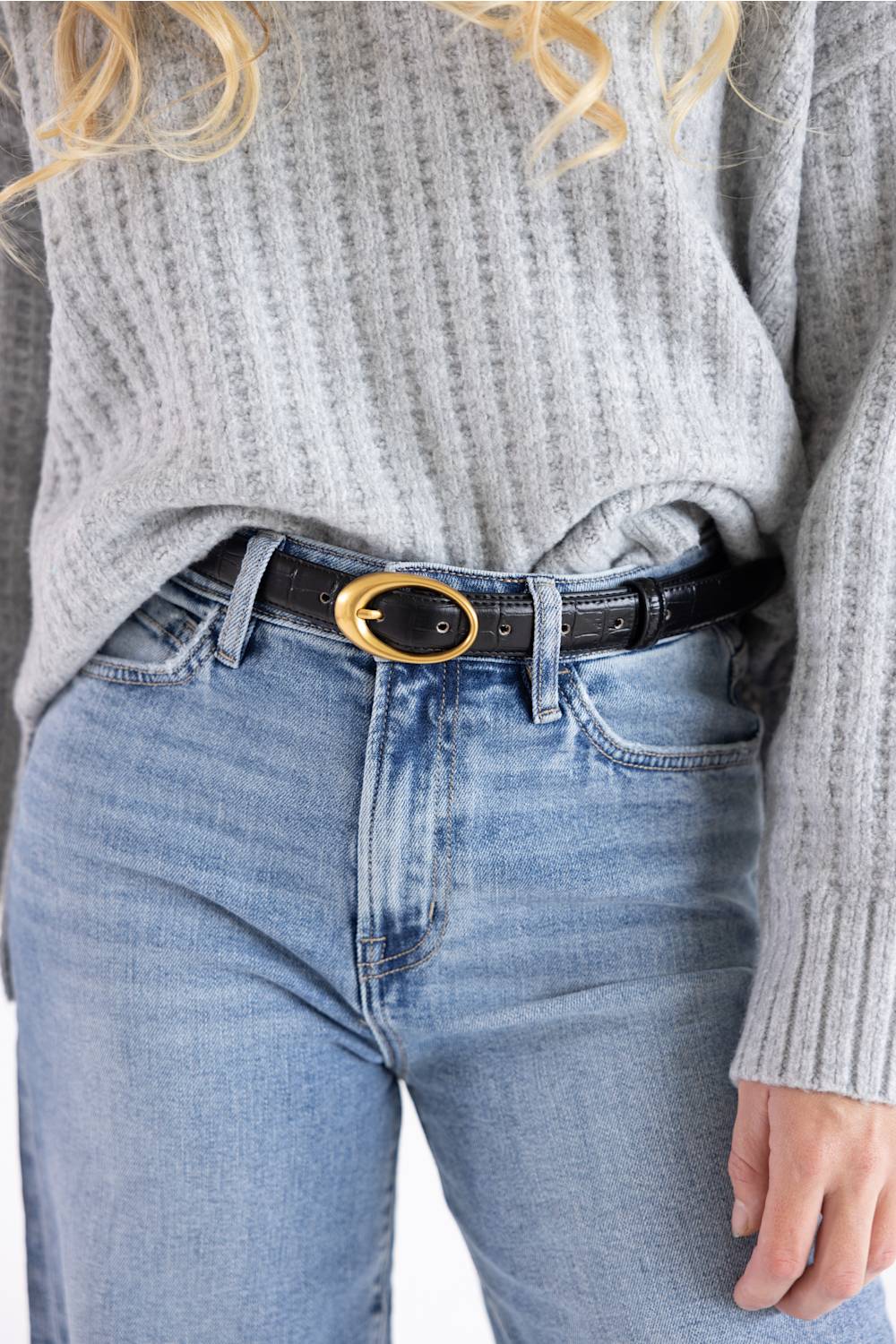 A close-up of a person wearing a black -leather belt with a large oval gold-tone buckle, styled with a gray ribbed knit sweater and light-wash blue jeans.