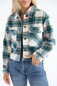 A woman wearing a cropped long-sleeve plaid button-up shirt jacket in teal green, white, and black, paired with blue jeans.