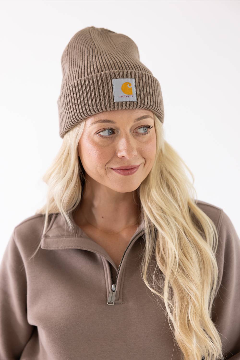 Blonde-haired woman smiling in a Carhartt Stone Brown cuffed rib knit beanie and coordinating brown half-zip sweatshirt.