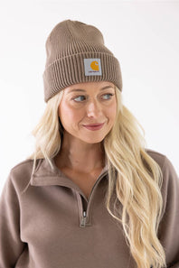 Blonde-haired woman smiling in a Carhartt Stone Brown cuffed rib knit beanie and coordinating brown half-zip sweatshirt.