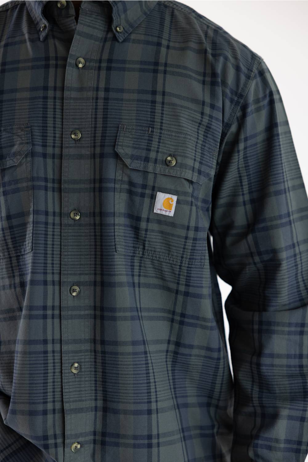 Close-up shot of a male model wearing the Carhartt Fort Loose Plaid Shirt in navy and green, highlighting the button-flap chest pocket, Carhartt logo patch, and dark buttons.