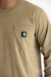 A close up of a man wearing a long sleeve light brown t-shirt with a black Carhartt patch on the upper left chest pocket.