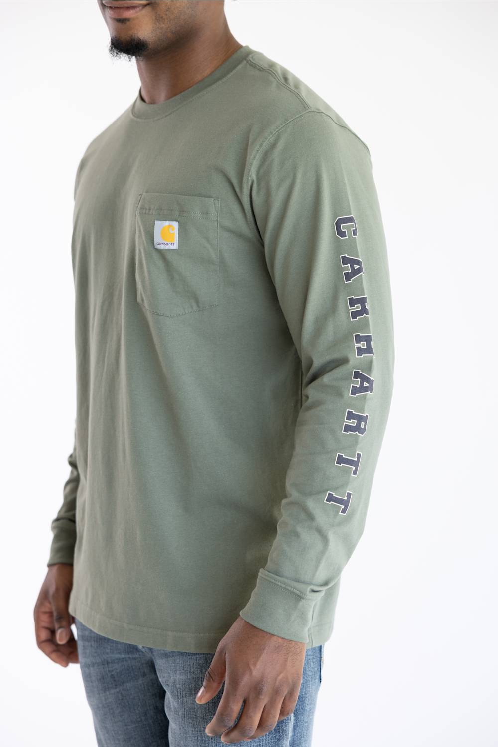A man wearing the Carhartt Irvine Relaxed Pointer Dog Long Sleeve T-Shirt for Men in Dusty Olive Green featuring a Carhartt logo down the left arm and a classic Carhartt patch logo with front pocket located on the upper left chest.