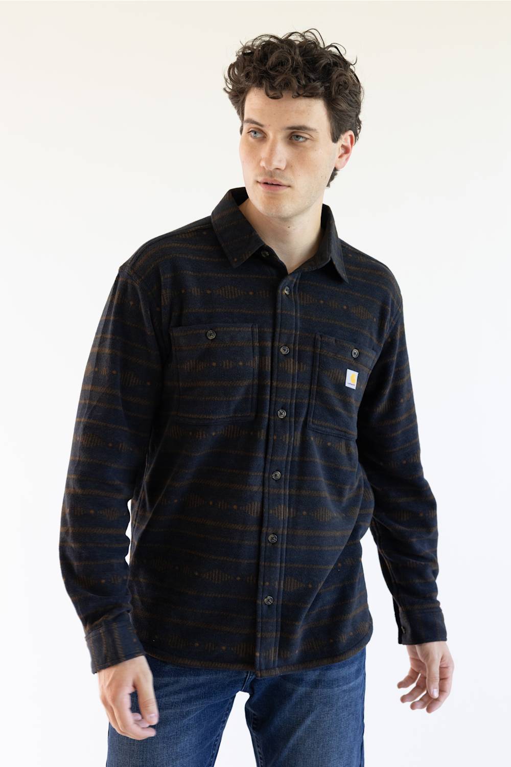 A man wearing a long-sleeve Carhartt plaid button-up shirt in navy and brown with thin orange stripes, paired with blue jeans, against a plain white background.