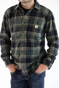 A man wearing a Carhartt long-sleeve flannel shirt in a green and black plaid pattern, paired with blue jeans, against a plain white background.