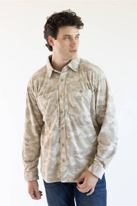 A man wearing a beige and light brown camouflage Carhartt long-sleeve button-up shirt with blue jeans, standing against a plain white background.