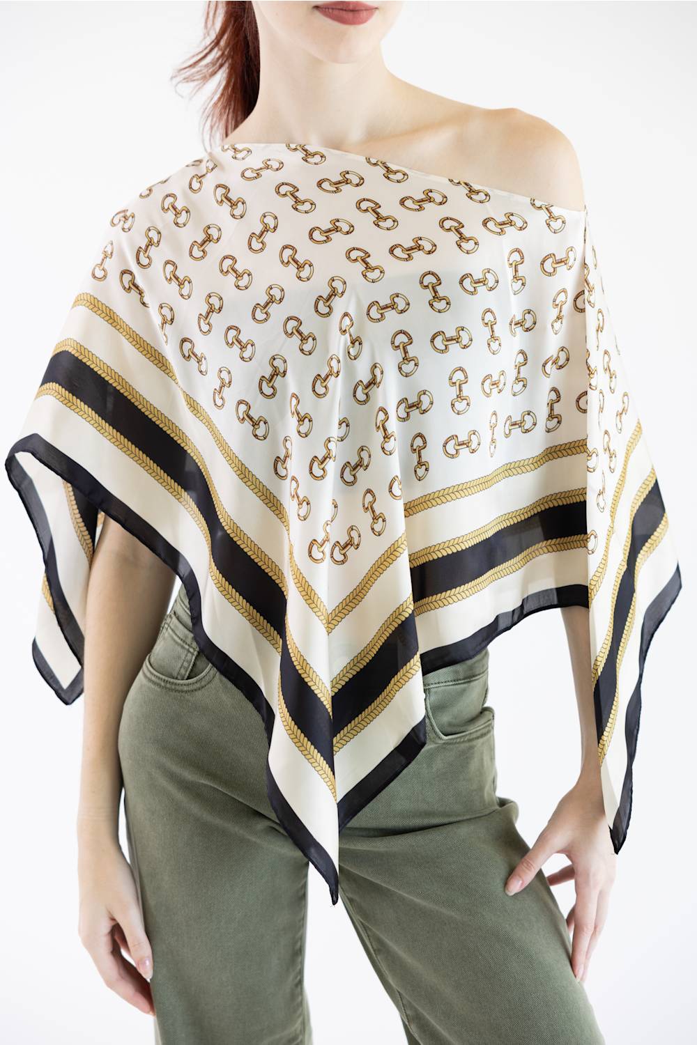 A woman wearing a white scarf top with a repeating brown interlocking logo pattern and black and gold striped borders, styled off-the-shoulder over an olive green top.