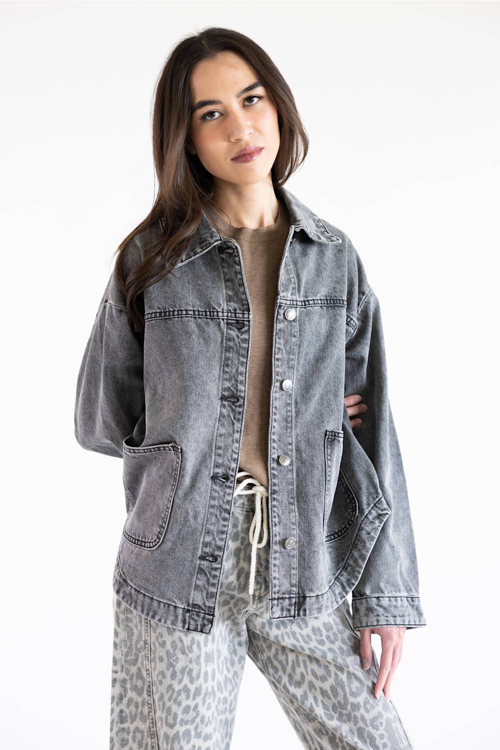 A woman modeling an oversized faded gray denim jacket over a beige top and gray leopard-print pants, photographed against a clean white studio background.