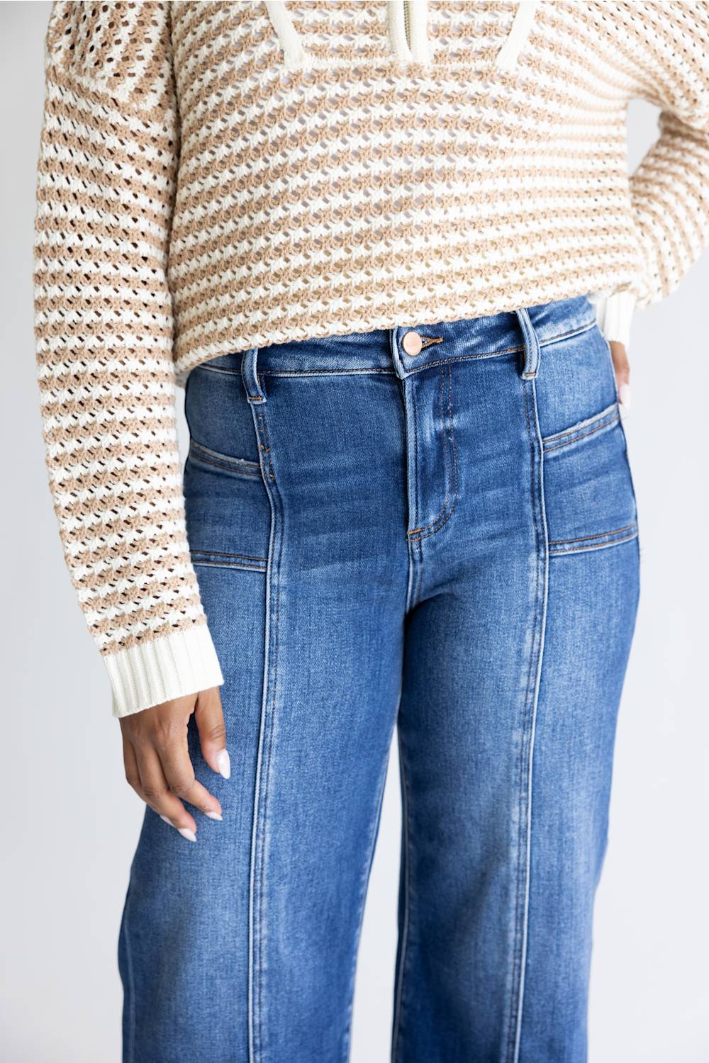A person wearing blue denim jeans and a cream and tan knit sweater, cropped from the chest down against a light gray studio background.
