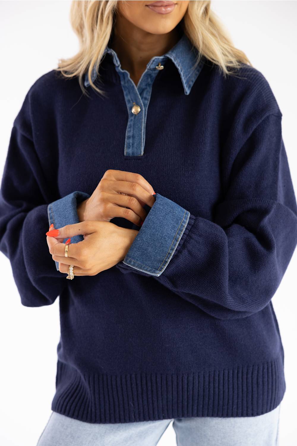 A woman wearing a navy blue knit sweater with contrasting blue denim collar, cuffs, and button placket, paired with light blue jeans against a white background.