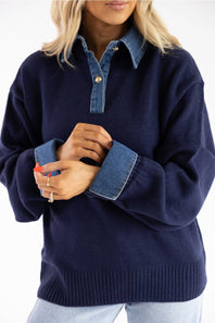 A woman wearing a navy blue knit sweater with contrasting blue denim collar, cuffs, and button placket, paired with light blue jeans against a white background.