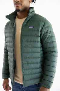 A man wearing a green Patagonia puffer jacket over a beige crewneck sweater and blue jeans, shown from the chin down against a plain white background.
