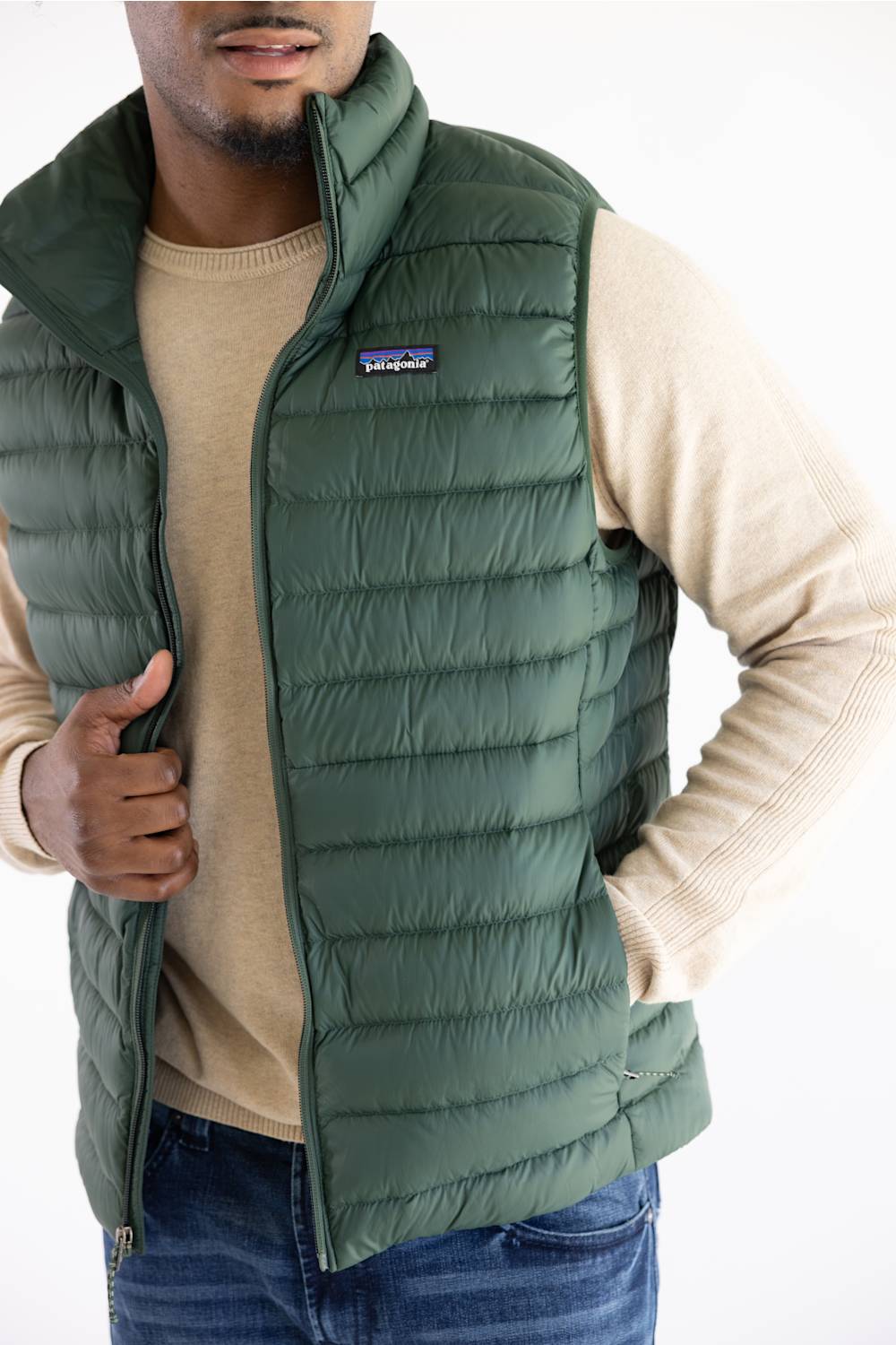 A man wearing a green Patagonia puffer vest over a beige sweater and blue jeans, shown from the chest down against a plain white background.