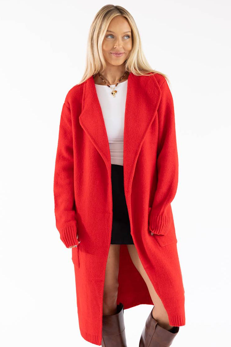 A woman modeling a bright red open-front knit cardigan over a white top, black skirt, and brown boots against a plain white studio background.