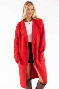 A woman modeling a bright red open-front knit cardigan over a white top, black skirt, and brown boots against a plain white studio background.