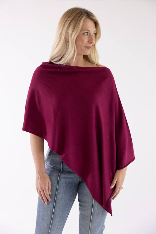 A woman wearing a draped, deep burgundy poncho-style top and light blue jeans, photographed against a plain white studio background.