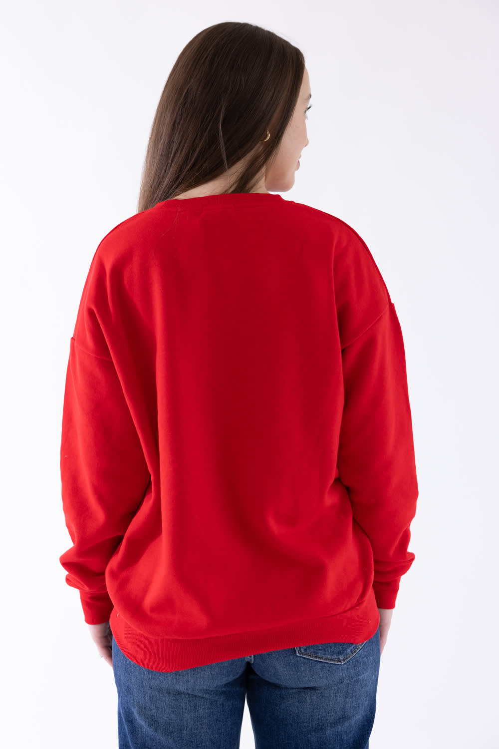 A woman viewed from the back wearing a bright red crewneck sweatshirt and blue jeans against a plain white studio background.